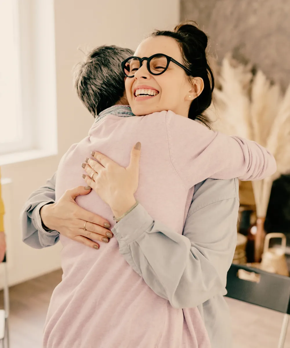 Two women share a warm hug, one smiling broadly over the other’s shoulder