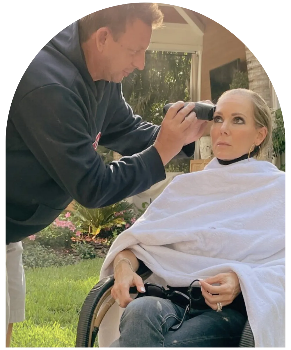 Tara Zuzga sitting outdoors in a salon cape while a man shaves her head during treatment